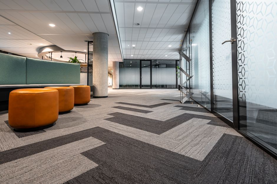 Spacious and modern office corridor featuring glass doors, geometric carpet patterns, and leather seating.